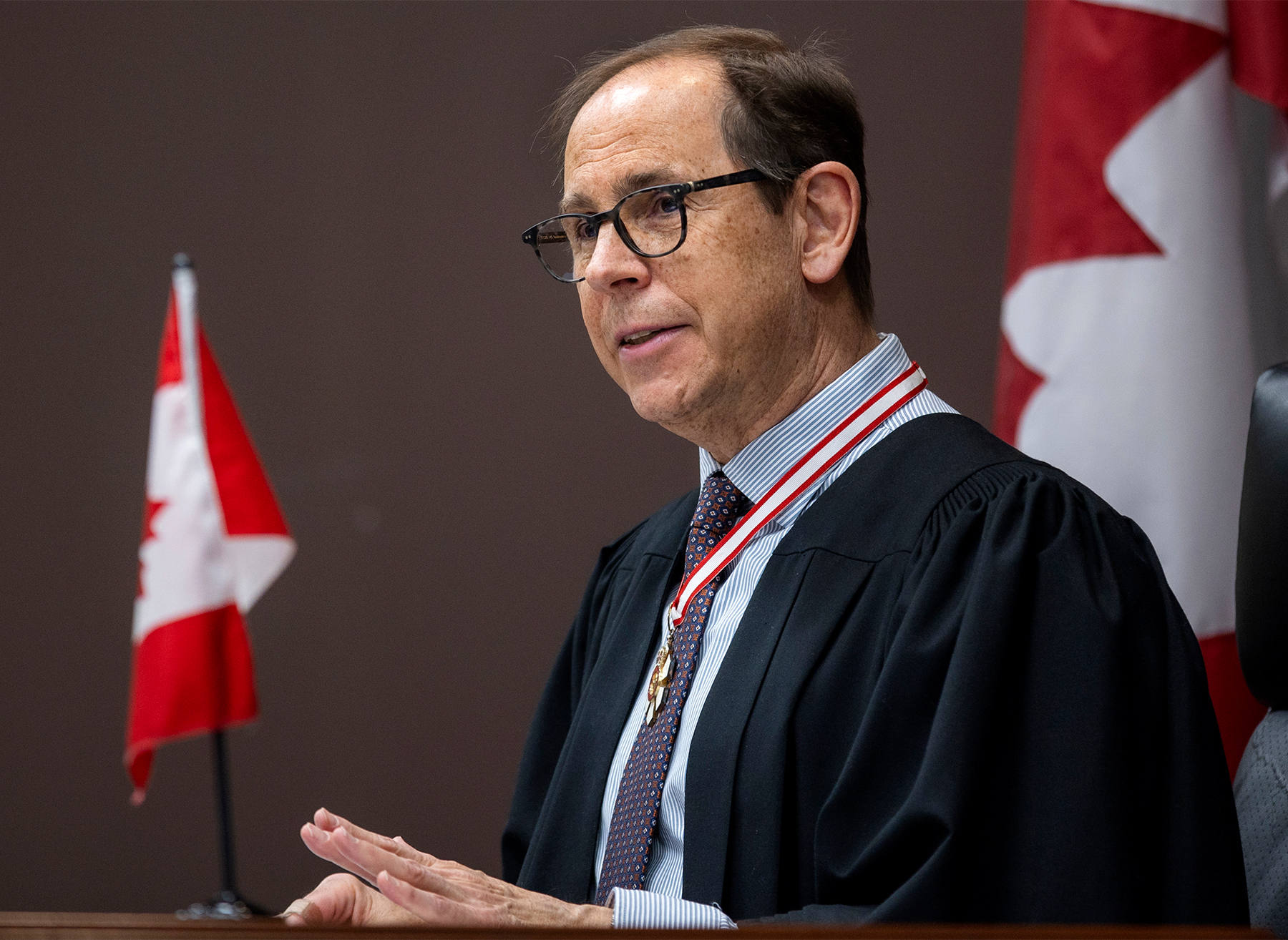 A white man wearing an order of Canada medallion and sitting at a desk with a Canadian flag on it addresses someone off-camera.