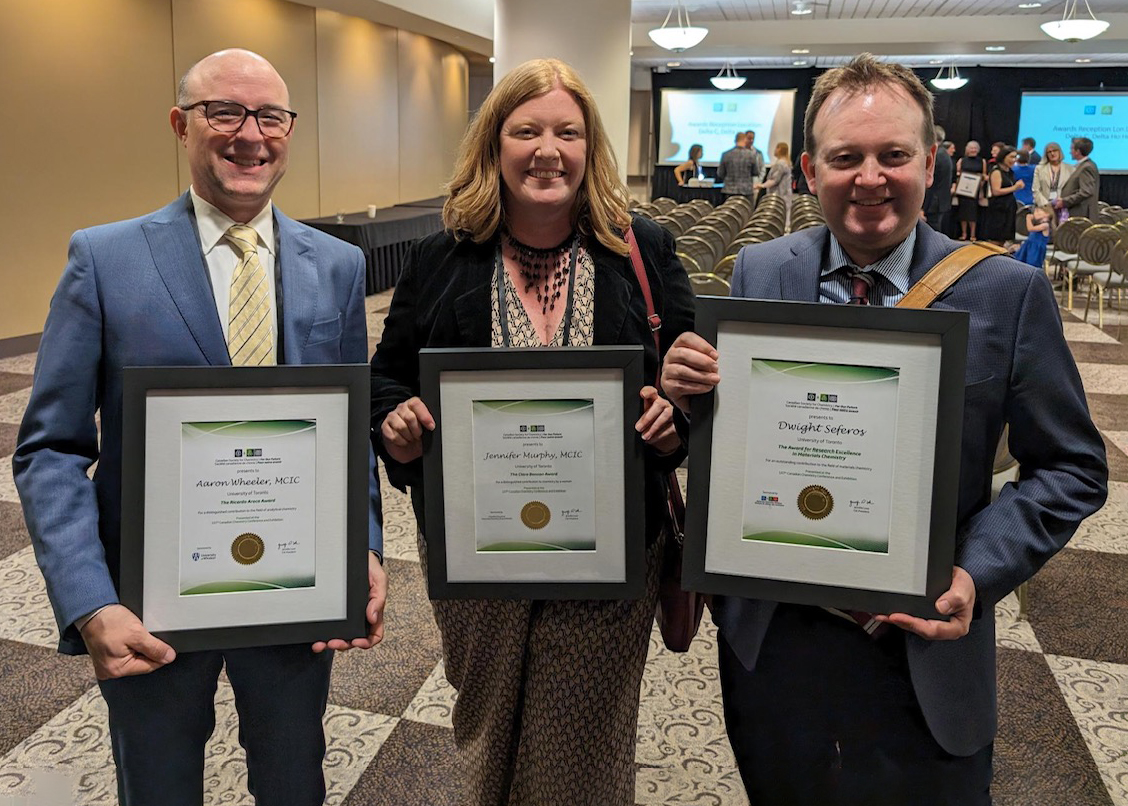 Three white adults holding prize certificates. There are two men flanking a woman with shoulder-length blonde hair.