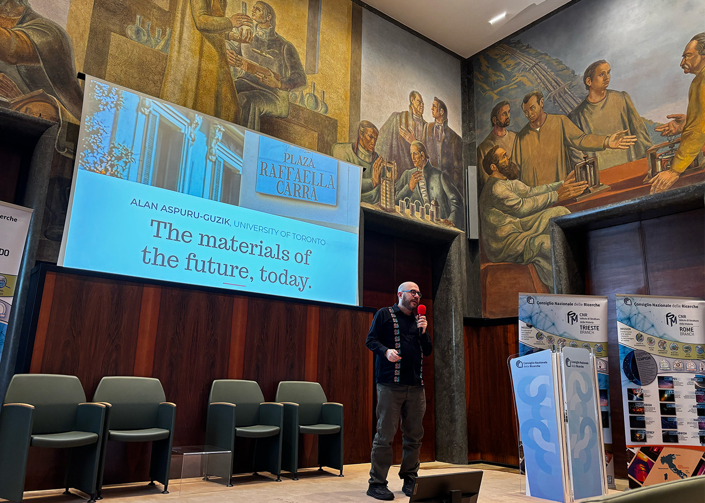 Chemist Alan Aspuru-Guzik addresses a crowd from a stage painted with frescos of Roman men.