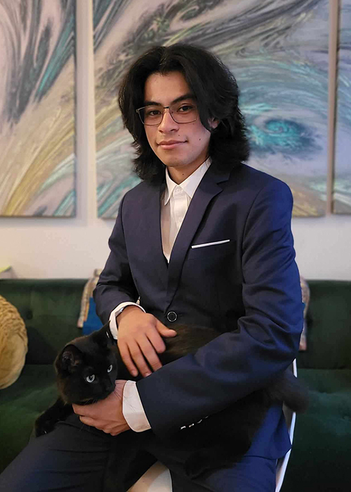 A El Salvadorean man with shoulder-length hair and a black cat sits on a stool in front of an abstract painting in blue and teal.