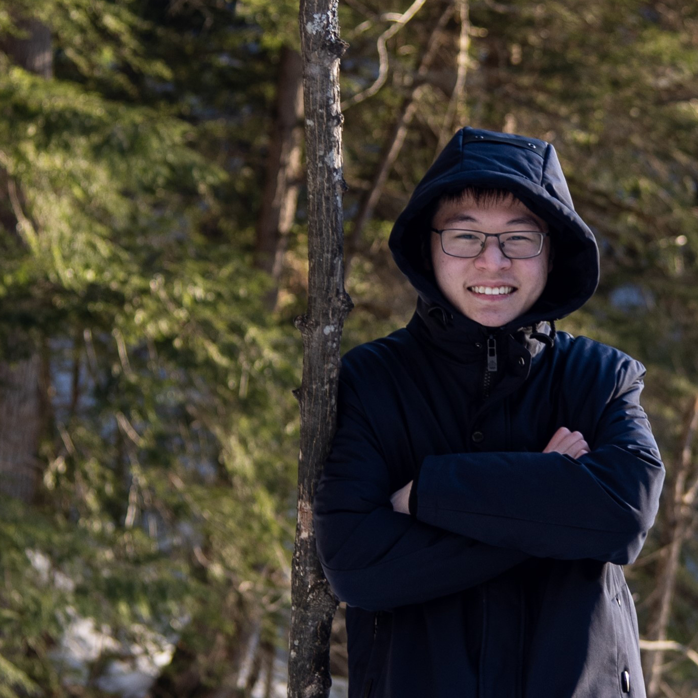 A young Asian man in a hooded jacket stands in a forest, grinning.
