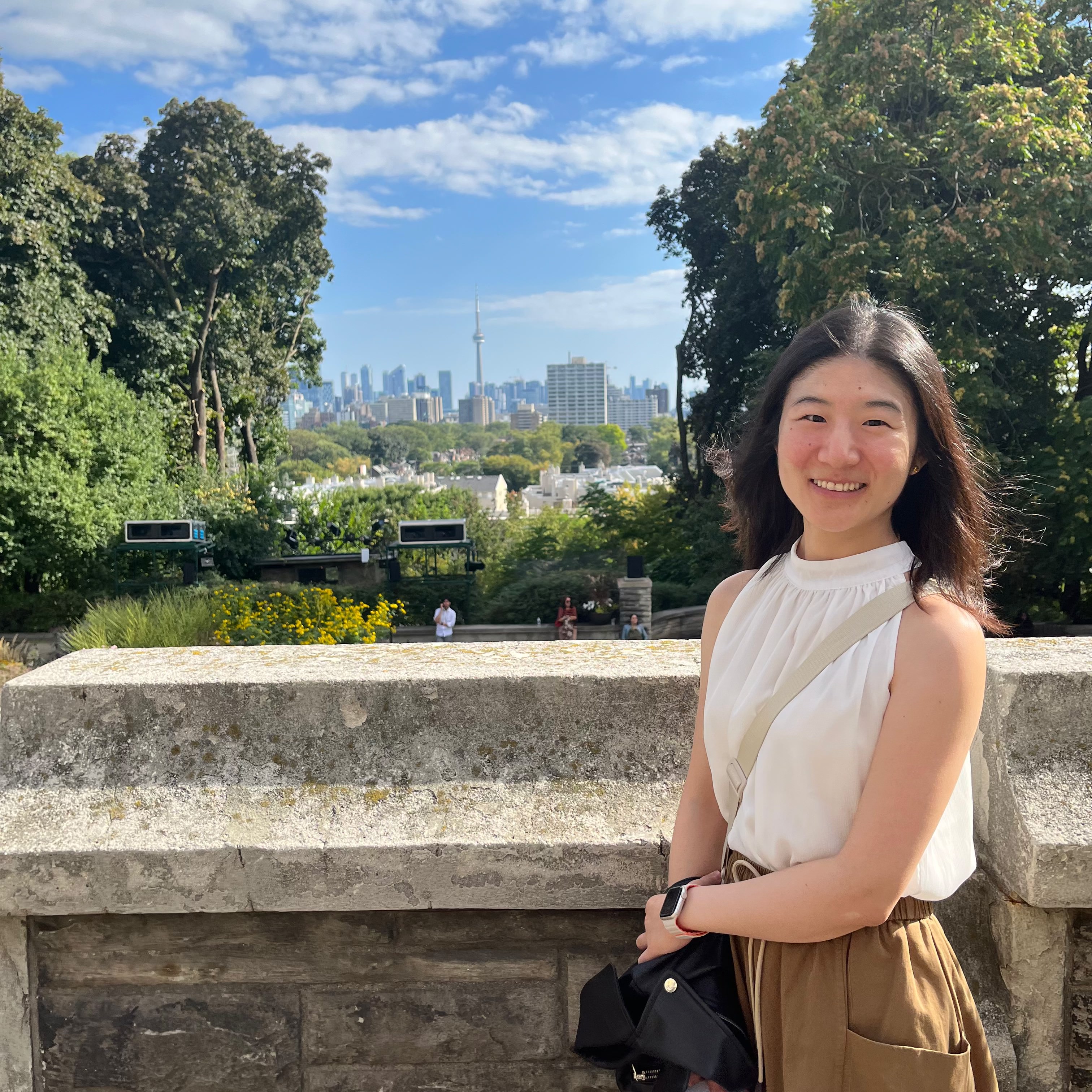 A young Asian woman in a white blouse, with long black hair, poses in front of a vista of downtown Toronto, complete with CN tower.
