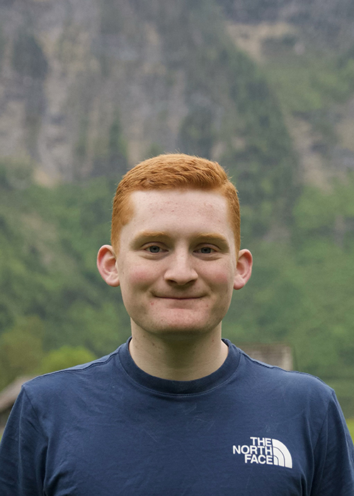 A young man with red hair in a blue shirt smiles at the camera. A blue and green mountain landscape is behind him.