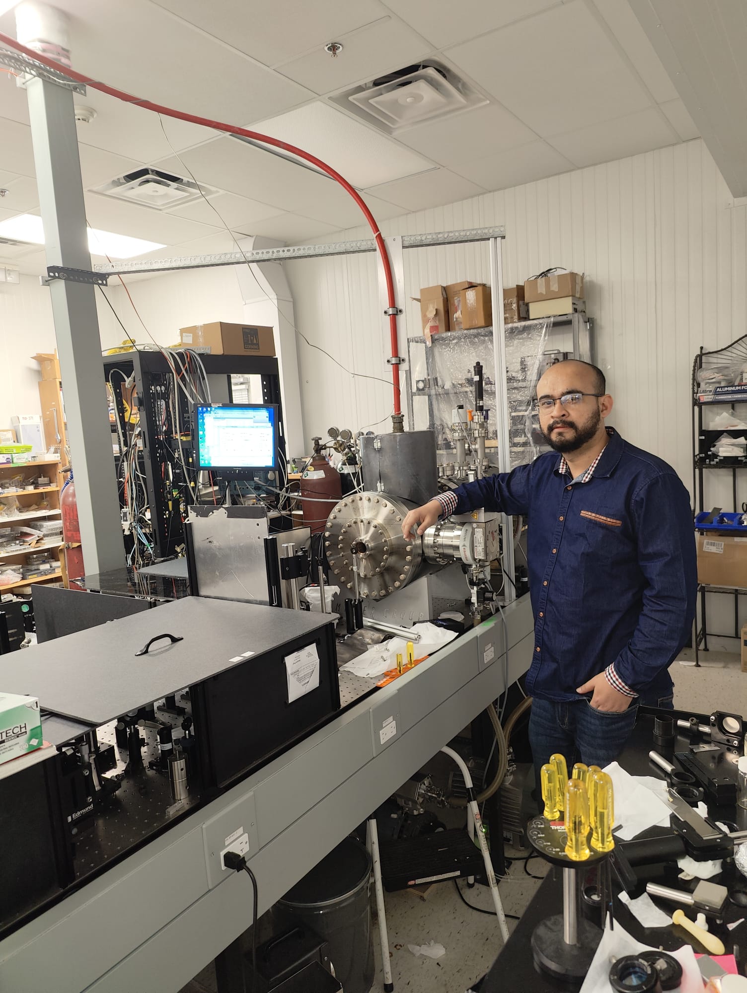 A man in glasses poses in a lab filled with equipment. He is wearing a blue shirt, and has black hair, moustache and beard.