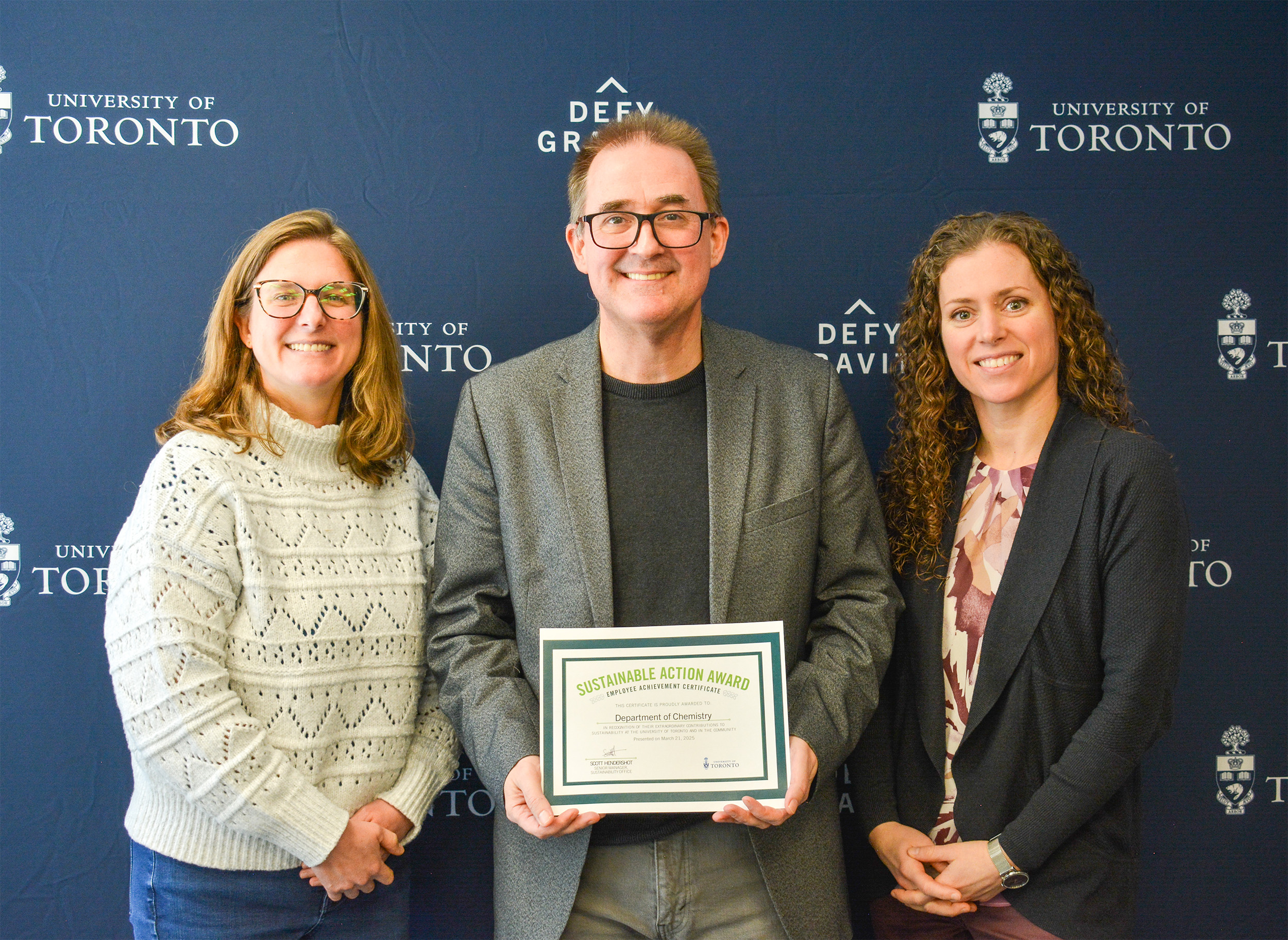 Three university professors; the middle professor, a man, holds an award plaque.