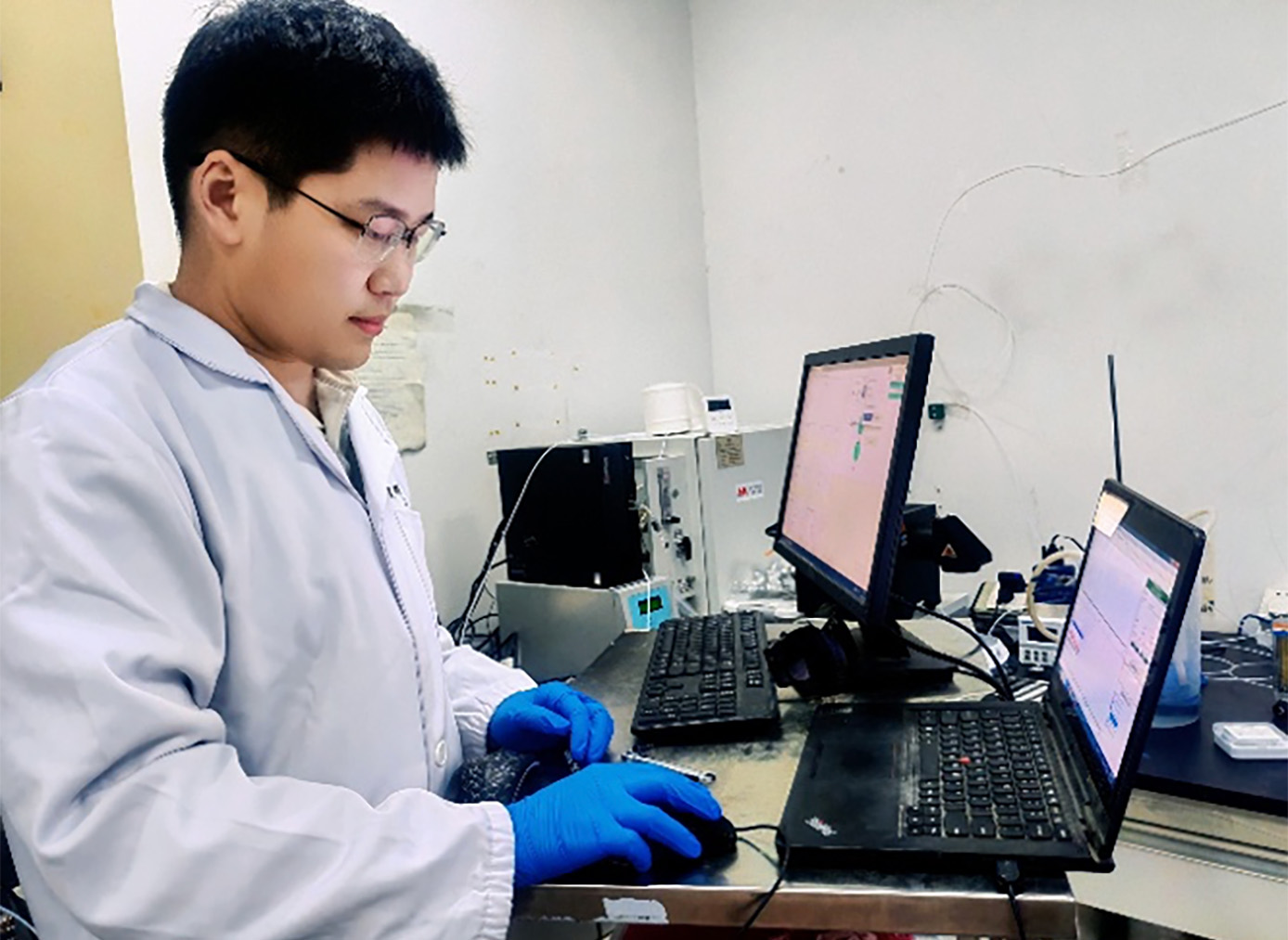 Wenqiang Qu works in lab gear in front of two laptops and lab equipment.