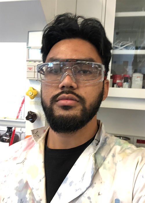 Closeup of an Arab man with moustache, beard and glasses, clad in a labcoat and looking with purpose into the camera.