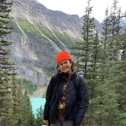 A smiling brown woman in a red hat standing in an evergreen forest smiles at the camera.