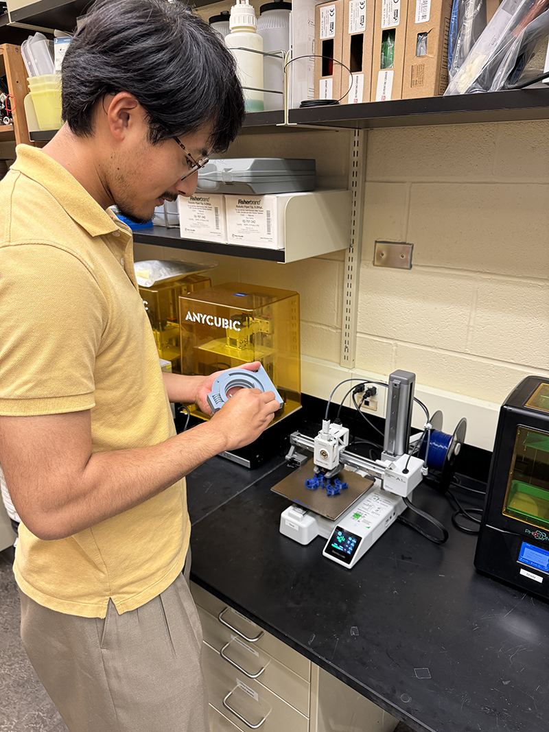 An Asian man  in a yellow shirt works on a fabricated part for a medical device.