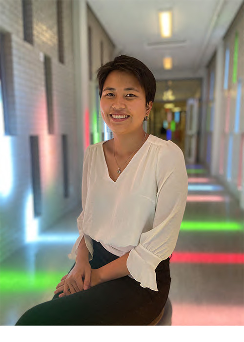 A South Asian woman with short hair sits at the fore of a multi-coloured corridor.