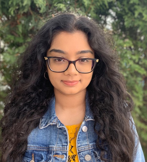 A young woman with long brown hair, jeans jacket and glasses smiles at the camera.