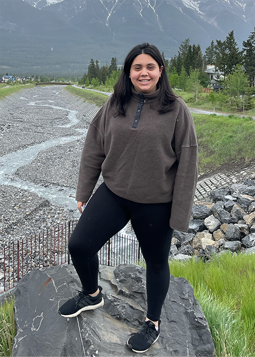 A young woman with long dark hair and athletic clothing poses in front of a mountain.