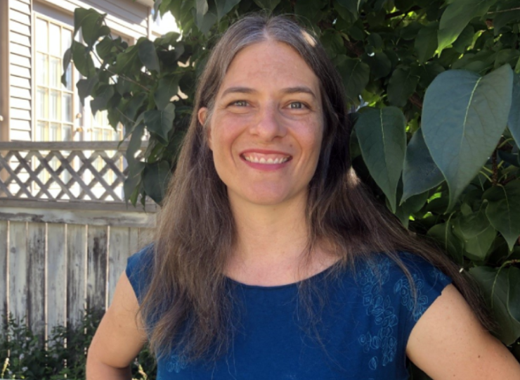 A white woman with long hair and a blue shirt stands, smiling, in front of a fence and a verdant green plant.