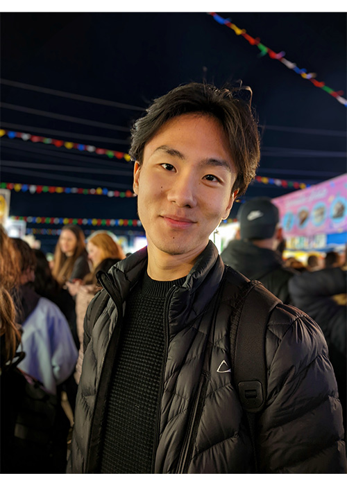 An Asian man with short hair and a black shirt and jacket smiles into the camera. A crowd at a fair is in the background behind him.