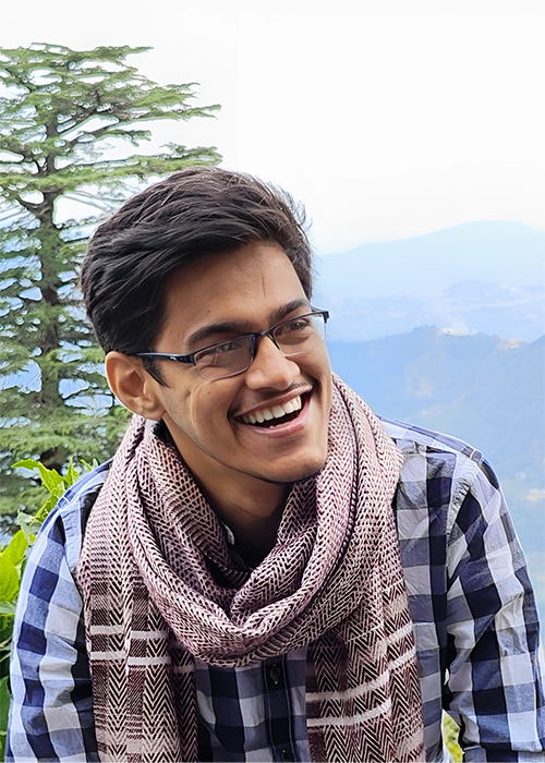 A South Asian man with glasses smiles in the foreground. Mountains and trees are behind him.