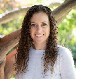 A white woman with shoulder-length curly brown hair stands in front of a tree