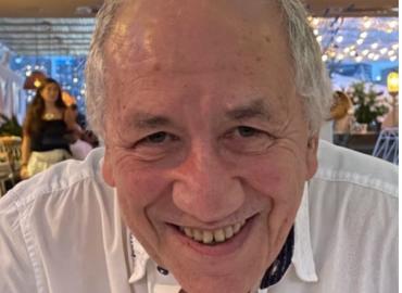 An older white man smiling and leaning toward the camera from a seated position at a table.