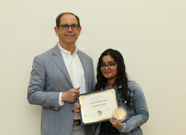 A white man on the left and a South Asian woman on the right smile at the camera. The woman is holding a certificate and and a medallion