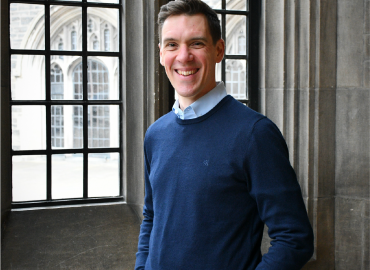 Head shot of a smiling white man standing in a blue shirt and sweater, standing in front of a tall window.