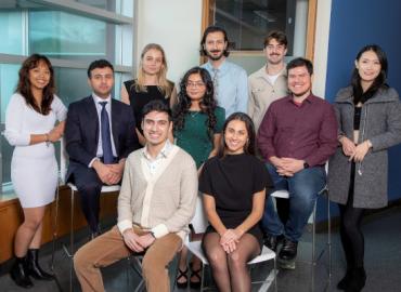 A group of ten people aged in their mid-twenties, some sitting and some standing. They are dressed in business casual attire, gathered in a group and smiling at the camera.