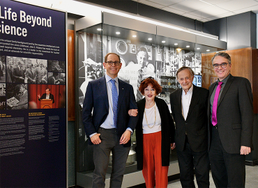 A man, woman and two more men gather with linked arms in front of a museum display, smiling at the camera.