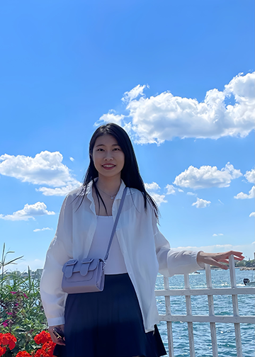 An Asian woman with long black hair stands in front of a river, leaning on a white fence next to a garden containing clusters of red flowers. The sky behind her is blue with white fluffy clouds.